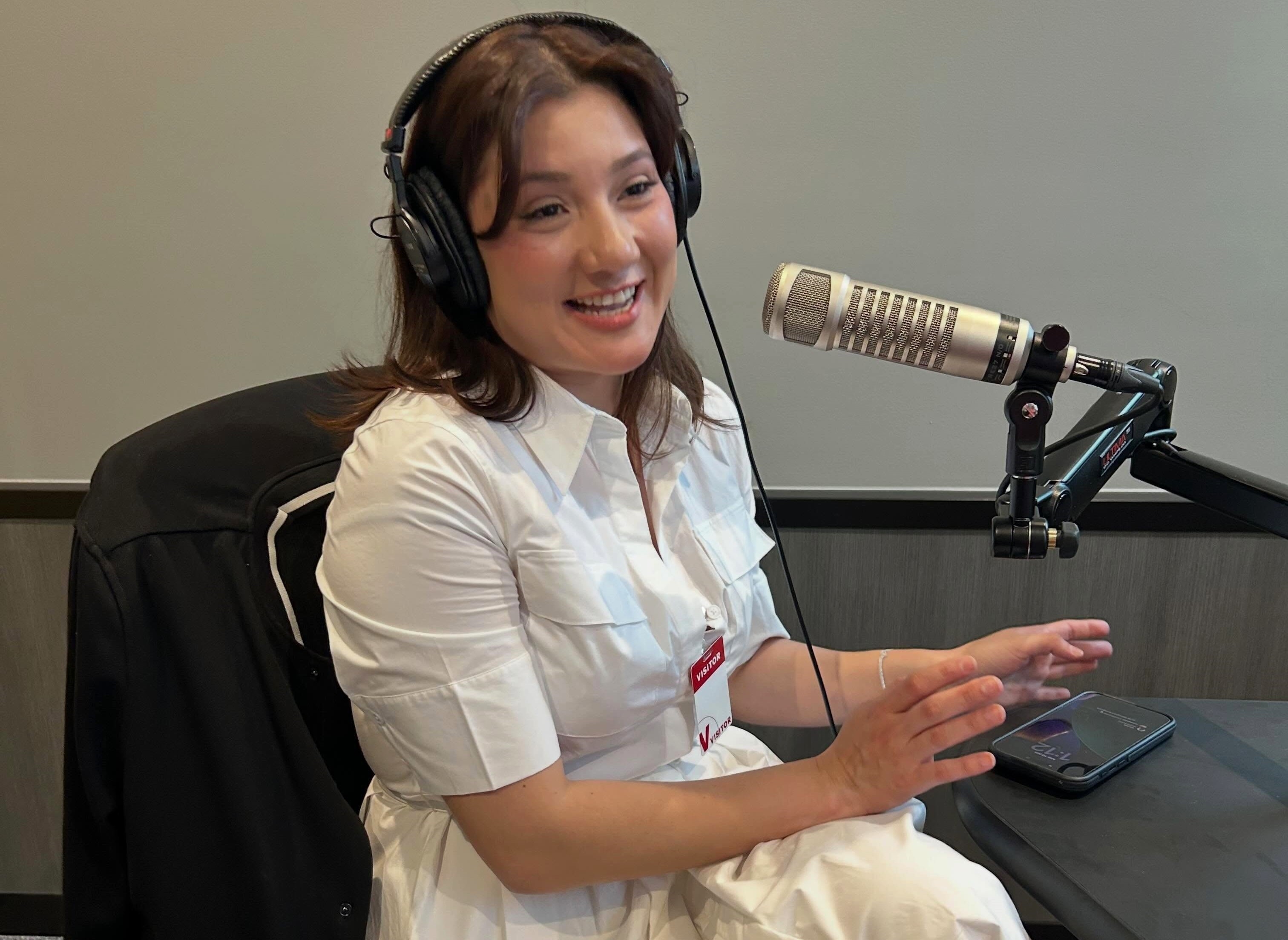 A smiling woman wearing headphones and speaking into a microphone while seated at a desk.